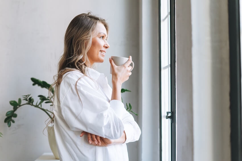 smiling woman holding a cup of coffee looking out of a window, anticipating the day ahead