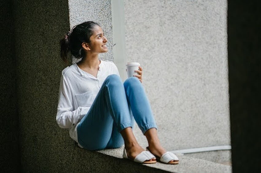 smiling woman sitting against a wall drinking coffee