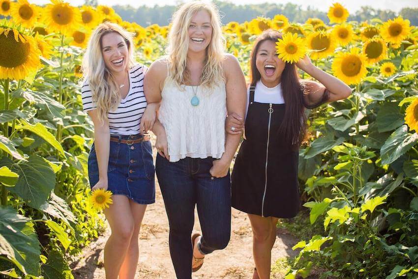 3 smiling women posing for the camera while standing in a sunflower field