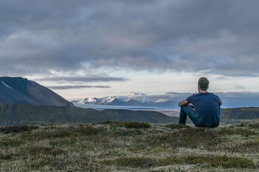 man sitting on the ground overlooking the top of mountains, lost in thought