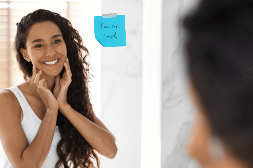 woman smiling at herself in the mirror with a sticky note that says You are loved!