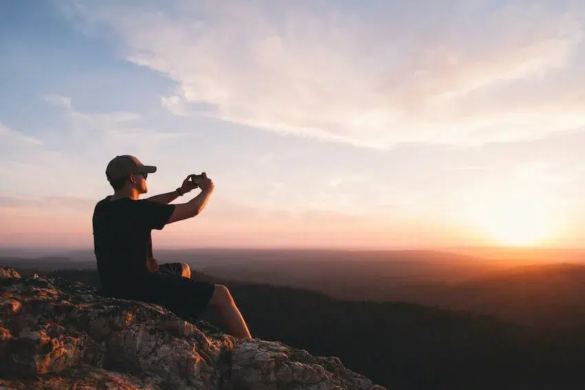 man sitting on top of a mountain taking a picture of the sunset