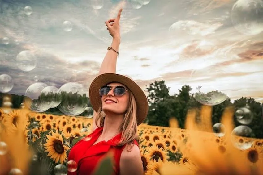 smiling woman with her right hand raised to the sky while standing in a field of sunflowers