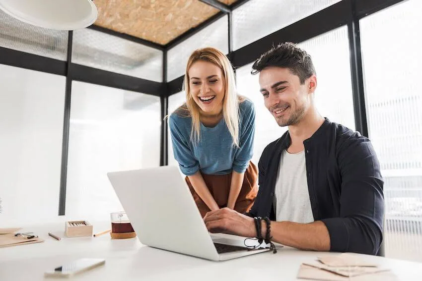 smiling woman overlooking a smiling man's shoulder as he types on his laptop