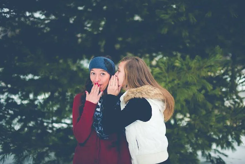 a female friend whispering in her friend's ear as she covers her mouth in surprise