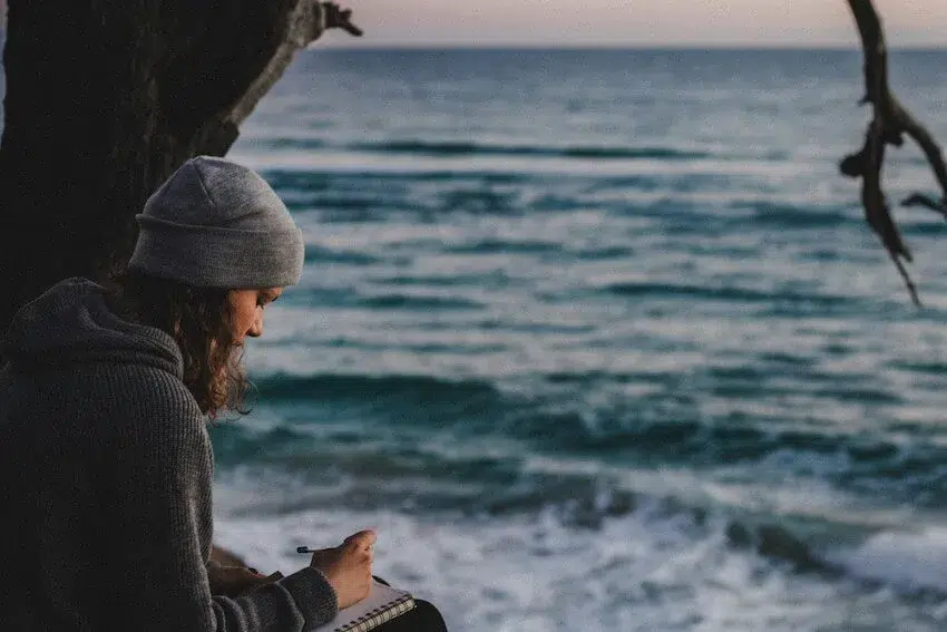 woman sitting next to the ocean writing in her journal