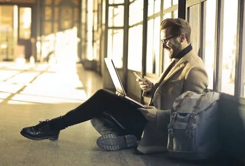smiling man sitting on the ground with his laptop, looking at his phone