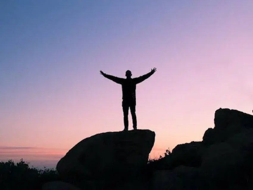 man standing on a large rock with his arms up at sunset