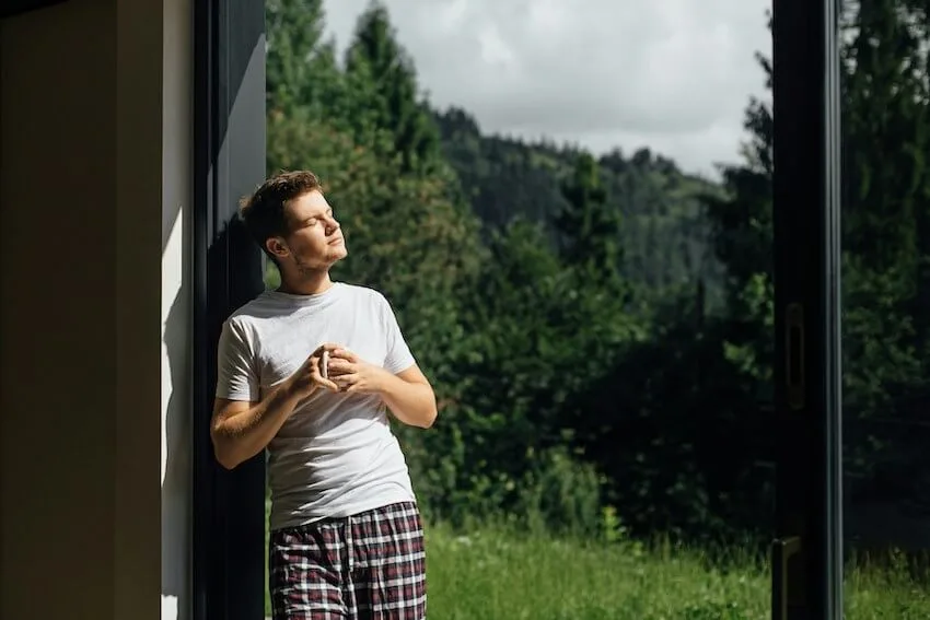 man in his pajamas holding a coffee mug while standing next to a window with the sun's rays streaming on his face
