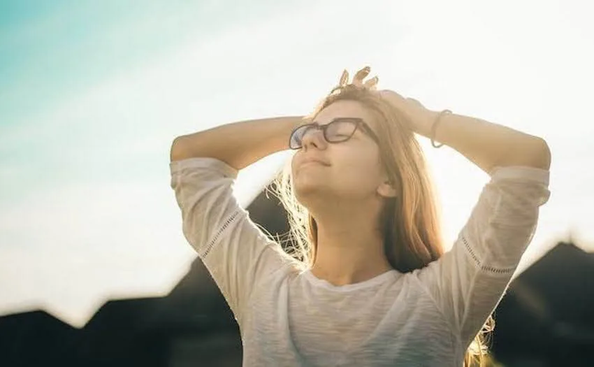 a smiling woman facing the sun with her eyes closed and her hands behind her head