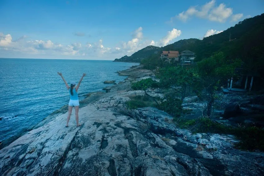 woman with her arms raised standing on top of a trail near an ocean