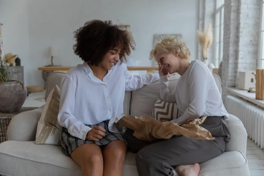 two friends sitting on a couch - one is smiling while she opens a gift while the other friend looks on