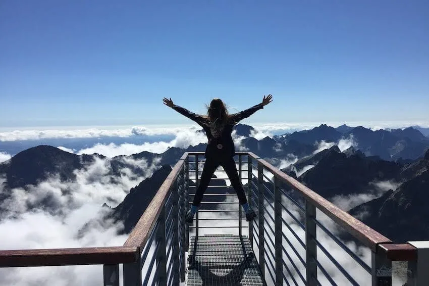 woman jumping with her arms and legs spread out at the edge of a path overlooking a mountain range